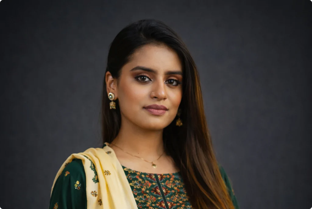 A woman with long dark hair, wearing a traditional embroidered dress with a green collar and gold jewelry, poses for a portrait inside a professional studio.