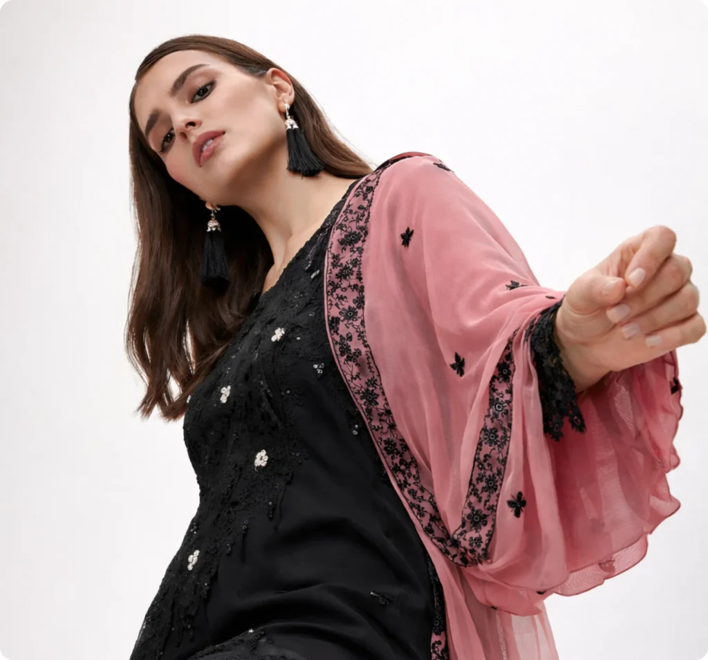 A woman posing in a black dress with a pink shawl draped over her shoulder inside a professional photo studio in Lahore.